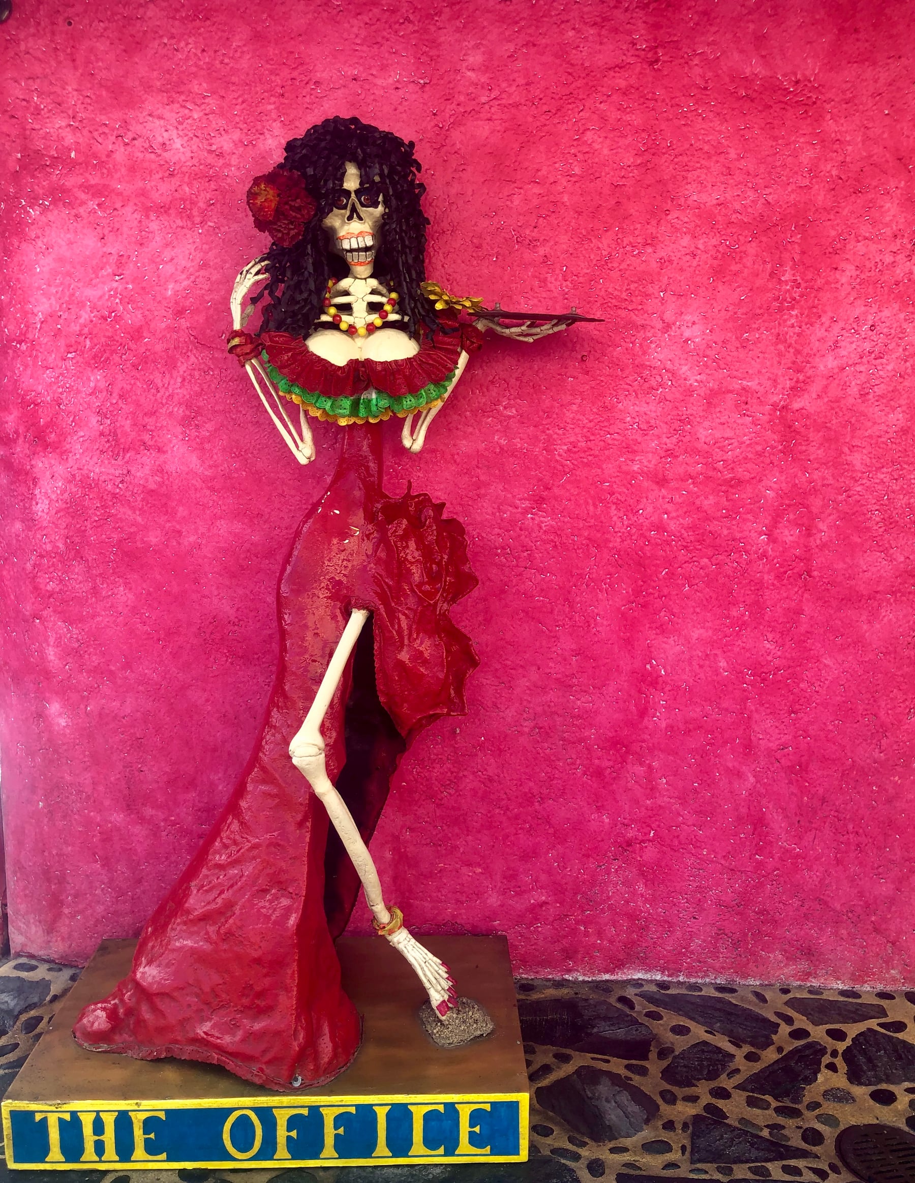 Decorative skeleton figure in a red dress against a bright pink wall at The Office on the Beach.